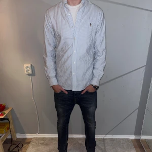 Randig skjorta från Ralph Lauren - Snygg ljusblå och vit randig skjorta från Ralph Lauren med klassisk krage och broderad logga på bröstet. Skjortan har lång ärm, knappar framtill och är tillverkad i mjuk bomull. Perfekt för en clean och stilren look. Modellen är 186 cm lång.