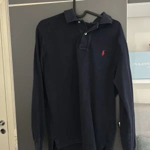 Mörkblå långärmad pikétröja Polo Ralph Lauren - Klassisk mörkblå långärmad pikétröja från Polo Ralph Lauren med röd broderad logga på bröstet. Tröjan har krage, knäppning med två knappar och är tillverkad i bomull. Perfekt för en stilren och avslappnad look. Den sitter något större än vad man tror så fråga först om längd Oså, inte använd alls mycket. Den är större där bak! Pris inte hugget i sten