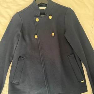 Mörkblå skepparkavaj Holly & Whyte XS - Stilren mörkblå skepparkavaj från Holly & Whyte med snygga guldknappar framtill och ståkrage. Jackan har raka fickor och en klassisk marin känsla. Perfekt för dig som vill ha en tidlös och trendig look.