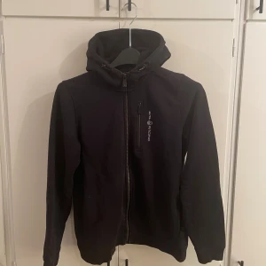 Svart fleecejacka Sail Racing - Svart fleecejacka från Sail Racing med huva och dragkedja hela vägen. Jackan har en bröstficka med dragkedja och logga på bröstet samt en broderad detalj på huvan. Mjuk insida i fleece och sportig look.