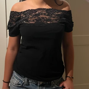 Svart offshoulder topp med spets - Snygg svart offshoulder topp med bred spets upptill som ger en romantisk vibe. Toppen är figurnära och har en ärmlös design med axlarna bara. Perfekt att matcha med jeans eller kjol för en trendig look.