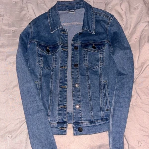 Klassisk blå jeansjacka - Säljer en klassisk blå jeansjacka med knappar framtill och två bröstfickor. Jackan har en rak passform, krage och långa ärmar. Perfekt att slänga över en hoodie eller t-shirt för en chill look. Strl xs från Vero Moda