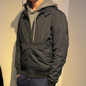 Svart bomberjacka med dragkedja - Snygg svart bomberjacka från j Lindeberg med ribbade muddar och hög krage med tryckknappar. Jackan har dragkedja framtill och två snedställda fickor med dragkedja. Tillverkad i ett slätt syntetmaterial som ger en clean och modern look.