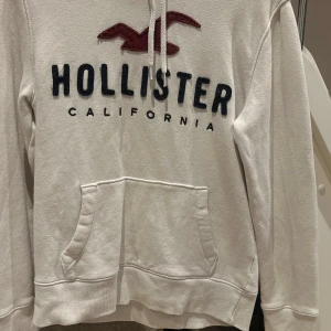 Vit hoodie från Hollister XS - Vit hoodie från Hollister med stor broderad logga och text på bröstet. Tröjan har huva med snörning, känguruficka och långa ärmar. Mjuk bomullskänsla och klassisk passform, perfekt för chill dagar.