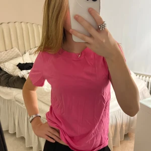 Rosa basic t-shirt i bomull från Kappahl  - Enkel och snygg rosa t-shirt i mjuk bomull. Klassisk rund halsringning och korta ärmar. Perfekt att styla med jeans eller shorts för en chill look. Färgen är riktigt poppig och ger outfiten ett lyft.