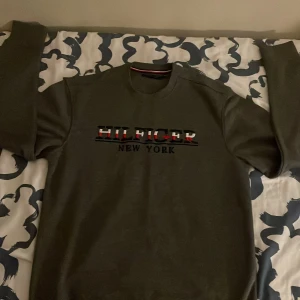 Mörkgrå Tommy Hilfiger sweatshirt - Säljer en mörkgrå sweatshirt från Tommy Hilfiger med broderad logga och texten 'HILFIGER NEW YORK' på bröstet. Tröjan har rund halsringning och långa ärmar. Perfekt för dig som gillar klassisk streetstyle. Passar även för L och M. Priset kan diskuteras 