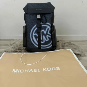 Michael Kors Ryggsäck - Helt Ny - Säljer en exklusiv och trendig ryggsäck från Michael Kors. Väskan är helt ny, oanvänd och kommer i oöppnad originalförpackning.  ✨ Detaljer: • Märke: Michael Kors • Färg: Svart med MK-monogram och stor vit logga framtill • Modell: Modern design med öppning och spänne framtill • Rymlig insida med flera fack för organisering • Sidofack med dragkedja för extra förvaring • Passar både till vardag, skola, jobb och resor  🎒 En snygg ryggsäck som kombinerar stil och funktionalitet.