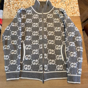 Grå kofta med GG-mönster från Gucci - Säljer en grå stickad kofta från Gucci med hel dragkedja och vit GG-logga över hela plagget. Koftan har ribbade muddar, hög krage och vita detaljer vid fickor och kanter. Tillverkad i 100% ull och perfekt för dig som vill sticka ut med lyxig streetstyle. Storlek L men passar M