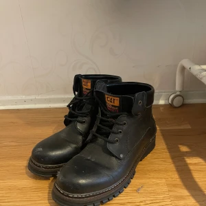 Svarta CAT boots i skinn - Tuffa svarta boots från CAT med grov sula och snörning. Skorna är tillverkade i slitstarkt skinn och har märkets logga i gult på baksidan och plösen. Perfekta för dig som gillar robust stil och vill ha något som sticker ut.