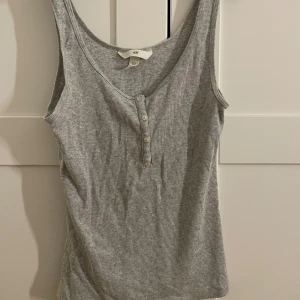Grå ribbad linnetopp från H&M - Säljer en enkel och stilren grå ribbad linnetopp från H&M i storlek S. Toppen har knappar framtill och är ärmlös med rundad halsringning. Perfekt att matcha med jeans eller kjol för en avslappnad look.