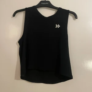 Svart ärmlös träningstopp från Hummel - Svart ärmlös träningstopp från Hummel med vitt logotryck på bröstet. Toppen är tillverkad i ett lätt och luftigt meshmaterial med snabbtorkande egenskaper. Modellen är croppad och har en loose passform, perfekt för gymmet eller löprundan.