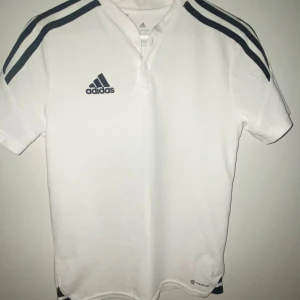 Vit pikétröja från Adidas med knappar - Vit pikétröja från Adidas med klassiska svarta ränder på axlarna och Adidas-logga på bröstet. Tröjan har korta ärmar, knappar vid halsen och är tillverkad i bomull. Perfekt för sport eller chill vardagsstil. Passar bra för ungdomar å vuxna, har inte används, säljer den för att det är inte riktigt min still. För 130 priset kan diskuteras