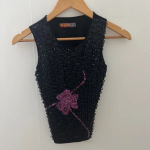 Svart paljettopp - Snygg svart ärmlös topp från utomlands med glittriga paljetter framtill och ett rosa blommotiv. Toppen har en croppad passform och slät baksida i stretchigt material. Perfekt för dig som vill sticka ut med lite extra bling. Aldrig använt den