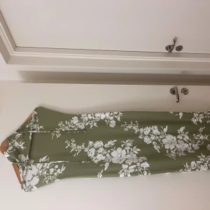 Grön blommig långklänning med volang - Långklänning i olivgrönt med stora vita blommor, ärmlös med smala axelband och knyt i nacken. Klänningen har en volangkant nertill och en luftig passform, perfekt för fester, semester . Materialet känns mjukt och svalt mot huden.