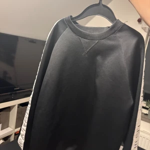 Svart sweatshirt från Les Deux - Svart sweatshirt från Les Deux med vita detaljer längs ärmarna där det står 'LES DEUX' tryckt. Tröjan har rund halsringning och långa ärmar. Perfekt för en clean och sportig look.