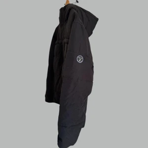 Svart pufferjacka från Peak Performance - Svart pufferjacka från Peak Performance med huva och diskret logga på ärmen. Jackan har dragkedja framtill, två bröstfickor med dragkedja och justerbara ärmslut. Perfekt för kalla dagar och riktigt snygg passform.