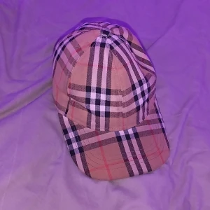 Rutig keps från Burberry - Säljer en klassisk keps från Burberry med ikoniskt rutigt mönster i beige, svart, vitt och rött. Kepsen har böjd skärm och justerbart spänne baktill i guldton. Tillverkad i bomull och polyester, perfekt för dig som vill ha en stilren accessoar.