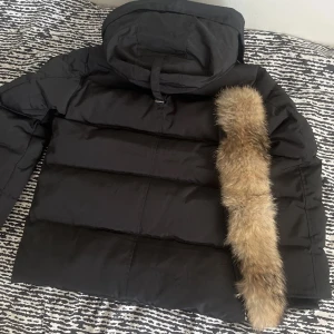 Svart dunjacka med päls från Canada Goose - Snygg svart dunjacka från Canada Goose med fluffig beige päls runt huvan. Jackan har lång ärm, diskret logga på ärmen och praktiska fickor framtill. Perfekt för kalla vinterdagar när du vill hålla stilen och värmen.