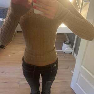 Beige kabelstickad tröja från GANT - Säljer en beige kabelstickad tröja från GANT med broderad logga på bröstet. Tröjan har en slim passform, ribbade muddar och är långärmad. Perfekt för lager-på-lager under höst och vinter. Supermjuk och klassisk stil!