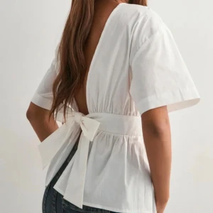 Vit blus med öppen rygg och rosett - ALDRIG ANVÄND LCH PRISLAPP KVAR Snygg vit blus i bomull med korta ärmar, markerad midja och stor rosett i ryggen. Toppen har en djup v-ringning bak och är lätt rynkad vid midjan för en feminin siluett. Perfekt för dig som vill ha en stilren men ändå unik look.