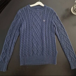 Blå kabelstickad tröja från Gant - Kabelstickad blå tröja från Gant med broderad logga på bröstet. Tröjan har ett klassiskt flätmönster och ribbade muddar vid ärmslut. Tröjan har inte använts jätte mycket och finns inga tecken på användning.