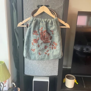 Broderad handväska H&M - Unik handväska från H&M i ljusblått bomullstyg med stora bruna andtag. Väskan har ett snyggt broderat blommönster i rostbrunt på framsidan och ett rymligt vinrött foder med innerficka och dragkedja. Perfekt för dig som vill sticka ut med en cool accessoar.
