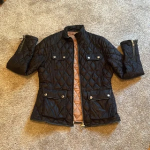 Svart quiltad jacka Barbour x Range Rover - Quiltad svart jacka från Barbour Range Rover Collection med snygga detaljer som silverfärgade knappar, dragkedja och fyra fickor framtill. Jackan har hög krage, lång ärm med dragkedja vid ärmslut och insida i champagnefärgad satin. Bara hängen är lite rostig annars inga defekter alls. Passar även S