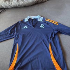 Real Madrid långärmad träningströja XL - Snygg långärmad Real Madrid träningströja från Adidas i mörkblått med orange detaljer och vit krage. Tröjan har halv dragkedja framtill, klubbmärke på bröstet och är tillverkad i lätt funktionsmaterial (Aeroready). Perfekt för fotbollsträning eller chill.