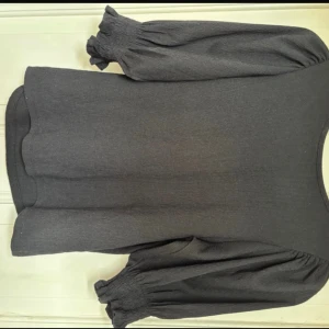 Svart blus med puffärm från Lindex - Svart blus från Lindex med puffiga trekvartsärmar och smock vid ärmslut. Blusen har en rund halsringning och är tillverkad i ett lätt, strukturerat tyg som ger en snygg och stilren look. Perfekt för dig som gillar trendiga detaljer och enkel design.