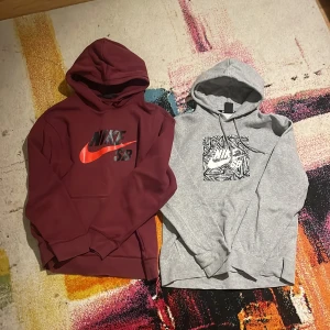 Nike SB hoodie vinröd & grå - Två snygga Nike SB hoodies, en i vinröd med röd logga och en i ljusgrå med svartvitt grafiskt tryck. Båda har huva, lång ärm och klassisk känguruficka framtill. Perfekt för chill eller streetwear.m-l