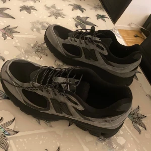 New Balance 2002R svarta/grå sneakers - Säljer ett par New Balance 2002R sneakers i svart och grå mocka och mesh. Skorna har en chunky sula, reflekterande detaljer och snabbsnörning. Perfekta för dig som gillar streetwear och vill ha en bekväm och snygg sneaker med sportig vibe.