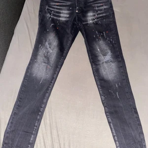 Svarta Dsquared2 jeans med färgstänk - Snygga svarta jeans från Dsquared2 med coola slitningar och färgstänk i vitt och rött. Jeansen har en smal passform och klassisk femficksdesign. Stor Dsquared2-patch baktill i rött och vitt. Perfekta för dig som vill sticka ut med en edgy look.