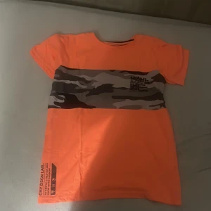 Orange camo t-shirt från Chapter Young - Cool orange t-shirt från Chapter Young i storlek 158/164. T-shirten har ett brett camouflagemönstrat band över bröstet och tryckdetaljer med text. Klassisk passform och korta ärmar, perfekt för en avslappnad streetstyle.