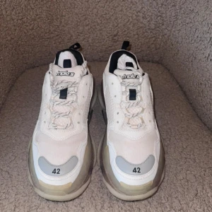 Balenciaga Triple S vita chunky sneakers - Säljer ett par Balenciaga Triple S sneakers i vitt med svarta och grå detaljer. Skorna har en chunky plattformssula, snörning och meshpaneler blandat med skinn. Ikonisk logga på plösen och storlek tryckt framtill. Perfekta för dig som gillar statement sneakers.