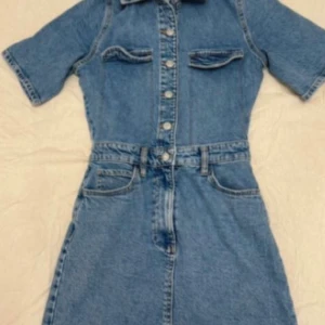 Kort jeansklänning med knappar - Snygg kort jeansklänning i blå denim med korta ärmar, krage och knappar hela vägen framtill. Klänningen har fickor på bröstet och i midjan samt bälteshällor. Perfekt för dig som gillar en cool och avslappnad stil.