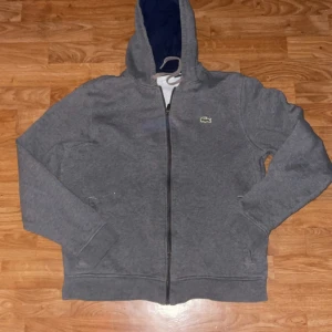 Grå hoodie från Lacoste Sport L - Säljer en grå hoodie från Lacoste Sport i storlek L. Tröjan har dragkedja, huva med snörning och klassisk krokodillogga på bröstet. Tillverkad i mjuk bomullsblandning med insida i fleece för extra komfort. Perfekt för chill dagar eller till träningen.