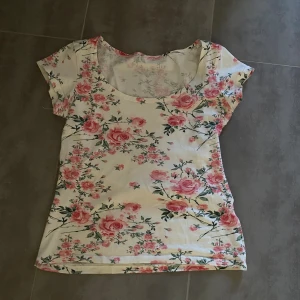Vit blommig topp med rosor - Söt vit topp med rosa blommor och gröna blad. Modellen har rund halsringning och korta ärmar.  Vet ej vart den är ifrån eller storlek men tror S
