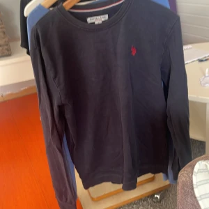 Svart långärmad tröja från US Polo Assn - Svart långärmad tröja från US Polo Assn i bomull med klassisk rund hals och röd broderad logga på bröstet. Enkel och stilren design som funkar till det mesta. Perfekt för dig som gillar en clean look.