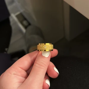 Bred guldfärgad ring med sten - Säljer en bred guldfärgad ring med snyggt graverat mönster och en liten glittrande sten i mitten från tilva. Ringen är i kirurgisk stål med en rundad insida vilket gör den behaglig och mjuk att ha på sig. Original pris ligger på 400 kronor.