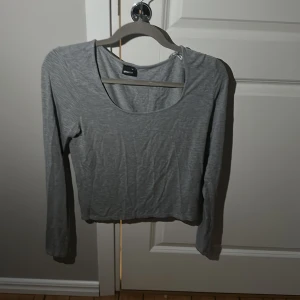 Grå långärmad topp  - En basic grå långärmad topp med rund halsringning och croppad passform. Tillverkad i mjukt bomullsmaterial, perfekt att styla med jeans eller kjol för en enkel och clean look.