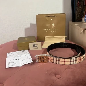 Burberry rutigt bälte med guldspänne - Snyggt bälte från Burberry med klassiskt rutigt mönster i beige, brunt och rött. Bältet har ett stort guldigt spänne med Burberrys TB-logga. Perfekt accessoar för att lyfta din outfit med en lyxig touch. Den är två sidig 