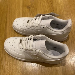 Nike Air Force 1 vita sneakers - Fräscha och klassiska Nike Air Force 1 sneakers i helvitt skinn med perforerad tå och platt sula. Snygga detaljer som metallbricka med 'AF1' på snörningen och ikonisk swoosh på sidan. Perfekt för dig som gillar stilrena och tidlösa sneakers. Anledning till varför jag säljer dem är för att dem är för stora. 
