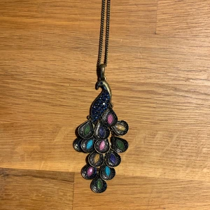 Två vintage halsband - Unikt halsband med en stor påfågelberlock i antikguld-look. Berlocken har glittriga detaljer i blått, grönt, lila, rosa och gult, och fjädrarna är droppformade med färgade stenar. Kedjan är lång och matchar den antika känslan. Perfekt statement-piece för dig som gillar färg och detaljer. Tyvärr saknar den en fjäder men det märks inte. Det andra halsbandet med lila/rosa stenar saknar två stenar men inget som syns på långt håll.
