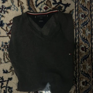 Mörkgrå v-ringad tröja Tommy Hilfiger - Mörkgrå v-ringad tröja från Tommy Hilfiger med diskret logga på bröstet. Tröjan är i bomull och har en klassisk passform med långa ärmar. Perfekt för dig som gillar stilrena och tidlösa plagg.