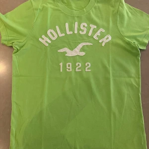 Grön Hollister t-shirt med tryck - Snygg grön t-shirt från Hollister med vit broderad logga, texten 'Hollister 1922' och klassisk rund hals. Skön passform och tillverkad i mjuk bomull, perfekt för en avslappnad stil.
