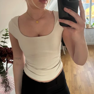 Beige croppad topp med rynkade sidor - Säljer en beige croppad topp med korta ärmar och rundad, djup urringning. Toppen har snygga rynkade detaljer i sidorna som ger en cool och smickrande passform. Perfekt att styla med både jeans och kjol.😍skriv vid frågor🥰
