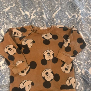Brun Mickey Mouse tröja Lindex - Säljer en brun långärmad tröja från Lindex med stort tryck av Mickey Mouse över hela. Tröjan har svarta och beige detaljer, rund hals och praktiska knappar på axeln för enkel påklädning. Perfekt för dig som älskar Disney och vill ha en skön vardagströja.