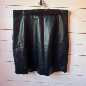 Svart skinnkjol i klassisk modell - Snygg svart kjol i skinnimitation med rak passform och enkel design. Kjolen är knälång och har en stilren look som passar till många olika outfits. Perfekt för dig som gillar en edgy och trendig stil.