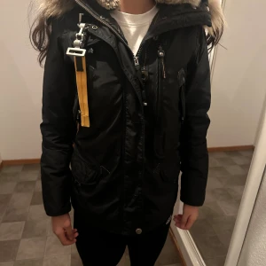 Svart Parajumpers parkajacka med päls - Säljer en svart parkajacka från Parajumpers med stor huva och fluffig päls runt kanten. Jackan har flera fickor, dragkedja och knappar framtill samt gula detaljer och patch på ärmen. Perfekt för kalla dagar och riktigt snygg streetstil.