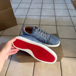 Blå mocka sneakers från Louboutin - Snygga blå sneakers i mocka från Louboutin med klassisk röd sula och vita kanter. Skorna har rund tå, snörning och diskreta detaljer i grått. Insidan är beige och de har en platt sula som ger en clean look. Perfekta för dig som gillar exklusiva märken och stilren design.
